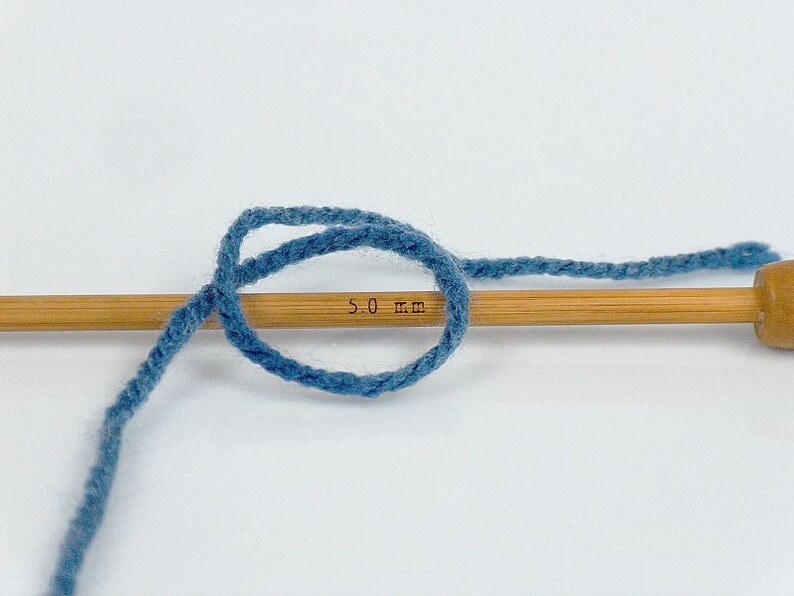 May include: A close-up of blue yarn wrapped around a wooden knitting needle with the text "5.0 mm" printed on the needle.