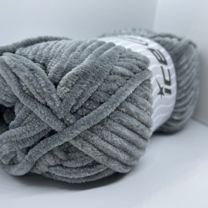 May include: A skein of grey yarn with a soft, fuzzy texture. The yarn is labelled "Ice Yarns" and is wrapped in a white label with a wavy design.
