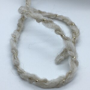 May include: A close-up of a beige yarn with gold glitter thread woven throughout. The yarn is twisted into a loop.