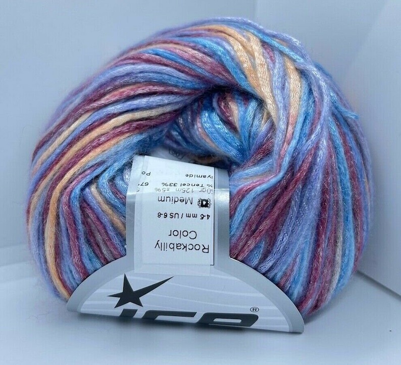 May include: A ball of variegated yarn featuring a blend of blue, pink, and peach tones. A white paper label is wrapped around the center, displaying the text Rockabilly Color, 4-6 mm / US 6-8, Medium, and 50g 125m 67% Tencel 33% Polyamide.