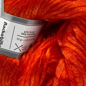 Puede incluir: Primer plano de una madeja de hilo naranja vibrante. La etiqueta dice "Rockabilly" y enumera el contenido de fibra como 67% Tencel y 33% Poliamida. La etiqueta tambi&eacute;n indica 50 gramos y 125 metros de hilo, con agujas de 4-5 mm.
