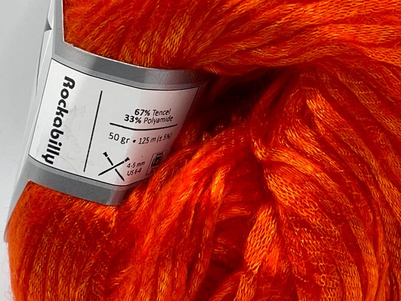 Puede incluir: Primer plano de una madeja de hilo naranja vibrante. La etiqueta dice "Rockabilly" y enumera el contenido de fibra como 67% Tencel y 33% Poliamida. La etiqueta tambi&eacute;n indica 50 gramos y 125 metros de hilo, con agujas de 4-5 mm.