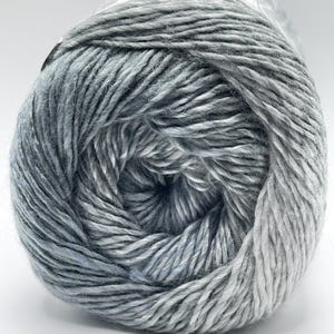 Può includere: Primo piano di un gomitolo di filato in tonalità grigie e blu. Il filato è avvolto strettamente, con fili visibili che creano una superficie strutturata. Il colore passa da un grigio chiaro a un grigio-blu più scuro, creando un effetto sfumato. Ideale per progetti di maglieria e uncinetto.