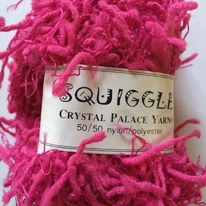 Puede incluir: Una bola de hilo rosa etiquetada "Squiggle Crystal Palace Yarns 50/50 nylon/polyester".