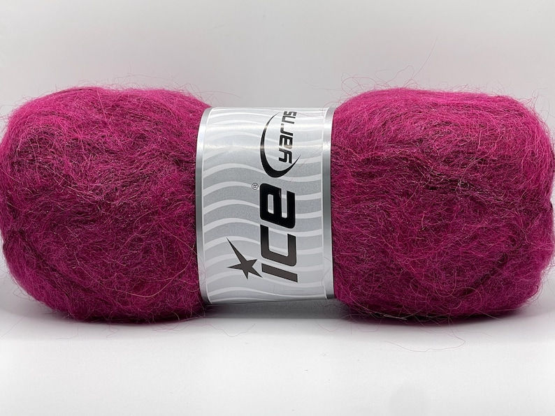 May include: A vibrant magenta yarn ball with a silver and white label. The label features the brand name "ICE" and a starburst design. The yarn appears soft and fluffy, ideal for knitting or crocheting projects.