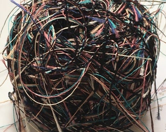 May include: A spherical ball of yarn composed of thin, multi-coloured strands in shades of black, teal, pink, and cream. The fibres are loosely wound, creating a textured, tangled appearance with numerous loose ends extending from the main structure.