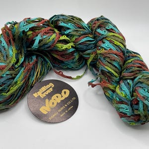 May include: A skein of Noro yarn in shades of teal, green, and brown. The yarn has a textured, ribbon-like appearance. A black tag with gold lettering reads "Knitting Fever Noro".