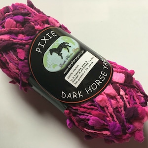 May include: A skein of pink and purple yarn with a label that reads "PIXIE Dark Horse Yarn" and includes information about the yarn, such as the fiber content, needle size, gauge, and yardage.