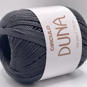 May include: A dark gray ball of yarn with a white label wrapped around it. The label reads "Círculo DUNA" and "186 yds / 170 m". The yarn is tightly wound and appears soft, suitable for knitting or crochet projects.