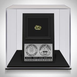 Puede incluir: Una muestra de Trinitita, un vidrio formado durante la prueba nuclear Trinity, exhibida en una vitrina transparente. La exhibición incluye una base negra y un marco negro que sostiene la Trinitita. El texto en la exhibición dice "TRINITITE - TRINITY ATOMIC BOMB GLASS".