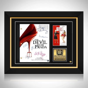 May include: Framed "The Devil Wears Prada" movie memorabilia. Includes a script page with signatures, two movie posters, and a gold-colored plaque. The frame is black with a gold inner border.