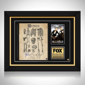 May include: Framed "Bones" TV show memorabilia. The frame has a black exterior with a gold interior border. The display includes a script page with anatomical drawings, a photo of the show's stars, and a Fox logo.