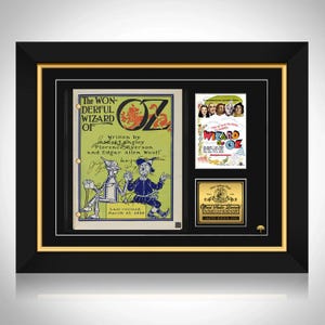 Puede incluir: Obra de arte enmarcada con una portada de libro vintage de "El Mago de Oz", un cartel de película y una placa dorada. La portada del libro tiene tonos verdes y amarillos con ilustraciones de personajes. El marco es negro con un borde interior dorado.