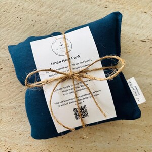May include: A rectangular dark blue linen heat pack wrapped in a white paper label tied with natural twine. The label features the text Linen Heat Pack, usage instructions, and an At Anchor logo. A small white fabric tag reads At Anchor Handmade.