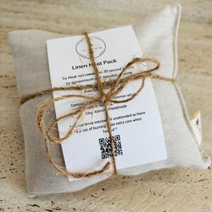 May include: A square, light beige linen heat pack tied with brown twine. A white instructional card is attached to the front, featuring the text Linen Heat Pack, heating instructions, a QR code, and the Etsy shop name AtAnchorHandmade.
