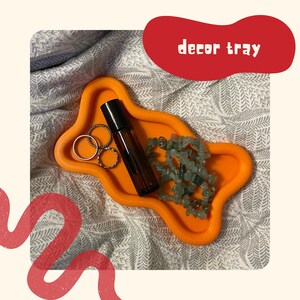 Op de afbeelding: Een oranje, golvend decoratief dienblad met een donker amberkleurige glazen rollerfles, drie zilverkleurige metalen ringen en een streng lichtgroene, getrommelde edelsteenkralen. De tekst decor tray staat in een wit, afgerond lettertype.