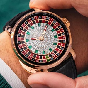May include: A rose gold wristwatch with a black leather band and a roulette wheel face. The face is decorated with diamonds and emeralds. The numbers 0, 29, 7, 28, 12, 35, 32, 6, 0, 32, 15, 19, 4, 21, 2, 25, 17, 34, 6, 27, 13, 36, 11, 30, 8, 23, 10, 5, 24, 16, 33, 1, 20, 14, 31, 9, 22, 18, 29, 7, 28, 12, 35, 3, 26 are visible on the face.