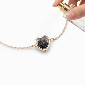 Puede incluir: Una pulsera de cadena dorada con un colgante en forma de corazón. El colgante tiene un marco dorado y contiene una esfera oscura y moteada. Una mano rocía la esfera con un frasco de perfume.