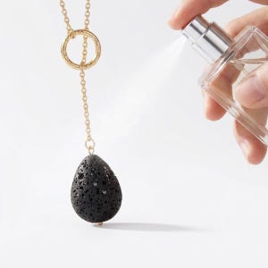 May include: A gold-toned lariat necklace featuring a delicate chain, a circular ring connector, and a single teardrop-shaped black porous lava stone pendant. The necklace is being misted by a clear glass spray bottle with a silver nozzle.