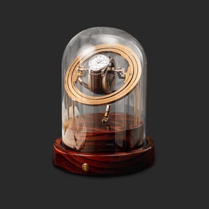 Op de afbeelding: Een horlogewinder met een glazen koepel, een houten basis en een goudkleurig roterend platform. De horlogewinder is ontworpen om een horloge opgewonden te houden en in goede staat te houden.