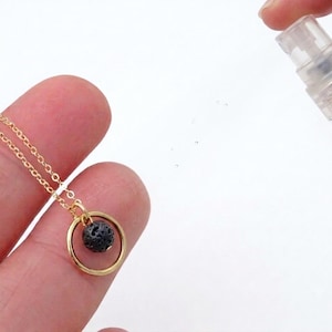 May include: A gold-toned chain necklace featuring a circular pendant with a small, porous black lava stone bead suspended in the center. A clear plastic spray bottle is positioned to the side, emitting a fine mist toward the necklace pendant.