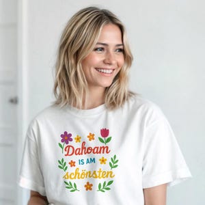 May include: White t-shirt with the text "Dahoam is am schönsten" in colorful floral lettering. The shirt is a crew neck style and is paired with light blue jeans. The overall aesthetic is casual and comfortable.