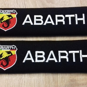Peut inclure: Deux épaulettes de ceinture de sécurité noires avec le texte "ABARTH" brodé en blanc et un logo de scorpion brodé en rouge, jaune et noir.