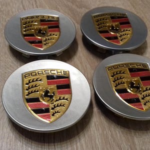 Peut inclure: Quatre enjoliveurs de roue argentés avec un logo Porsche noir et rouge au centre. Le logo est doré et comporte un blason avec un cheval au centre.