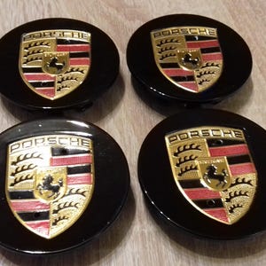 Peut inclure: Quatre enjoliveurs de roue noirs avec le logo Porsche en or et rouge. Le logo représente un cheval et un blason avec des feuilles.