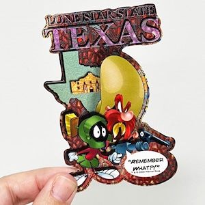 Könnte beinhalten: Ein bunter Magnet in Form des Bundesstaates Texas mit den Worten "Lone Star State" und "Texas" oben. Der Magnet zeigt Zeichentrickfiguren aus den Looney Tunes, darunter Yosemite Sam und Marvin der Marsmensch. Der Text "Remember What?" ist ebenfalls auf dem Magneten enthalten.
