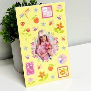 May include: A yellow sticker sheet featuring a Barbie doll in a pink outfit holding flowers. The sheet is decorated with floral and insect illustrations, including dragonflies, bees, and flowers. The word "Barbie" is printed on the sheet.