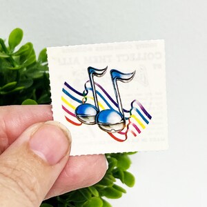 May include: A small, square sticker featuring two blue musical notes with rainbow accents. The notes are set against a white background with a scalloped edge. The sticker has a vintage aesthetic.