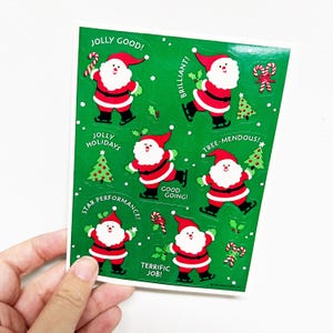 Peut inclure: Une planche d'autocollants de Noël avec des illustrations du Père Noël sur fond vert. Les autocollants comportent des phrases comme "Jolly Good!" et "Tree-mendous!" ainsi que des cannes en sucre et des sapins.