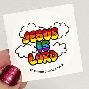 May include: A round sticker with the text "JESUS IS LORD" in rainbow colors, outlined in black. The design includes white cloud shapes and the copyright information "Sonrise Creations 1983".