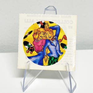May include: A square, vintage-style print featuring a cartoon couple dancing in front of a yellow circle with vinyl records and musical notes. The man wears a blue suit and the woman a pink top. The print is displayed on a clear acrylic stand.