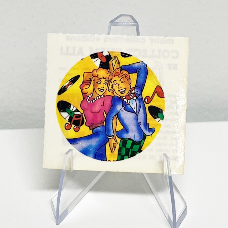 May include: A square, vintage-style print featuring a cartoon couple dancing in front of a yellow circle with vinyl records and musical notes. The man wears a blue suit and the woman a pink top. The print is displayed on a clear acrylic stand.