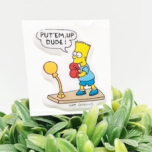 Könnte beinhalten: Ein klarer Aufkleber mit Bart Simpson in Boxerkleidung, mit roten Handschuhen und blauen Shorts, der auf einer Holzplattform steht. Eine Sprechblase über ihm lautet "PUT'EM UP DUDE!". Der Aufkleber befindet sich vor einem Hintergrund aus grünem Laub.
