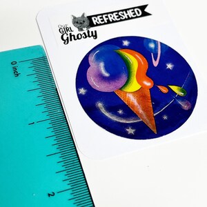 May include: A sticker with a vibrant ice cream cone design. The scoop is a rainbow of colours, and the cone is brown, set against a dark blue backdrop with stars. The sticker includes the text "Our Girl Ghosty" and "Refreshed".