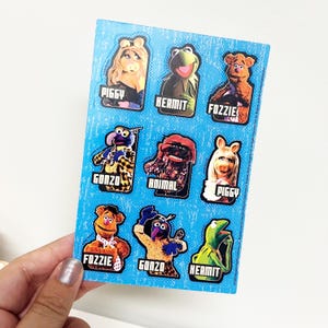 May include: A sheet of stickers featuring various Muppets characters, including Miss Piggy, Kermit, Fozzie Bear, and Gonzo. The stickers are on a blue background, with each character's name printed below their image.