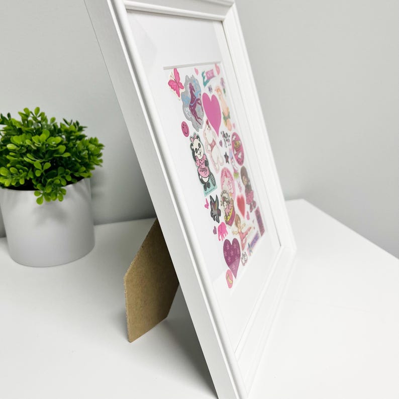 Puede incluir: Obra de arte enmarcada en blanco con un collage de pegatinas coloridas. Las pegatinas incluyen corazones, animales y otros dise&ntilde;os caprichosos. La obra de arte se exhibe sobre una superficie blanca con una peque&ntilde;a planta en maceta en el fondo.