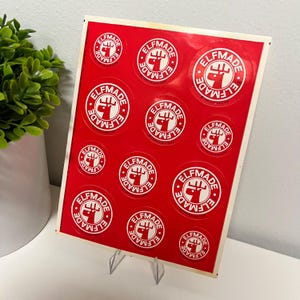 May include: A sheet of red stickers featuring the text "ELFMADE" in a white circle with a red graphic of a fist holding a moose antler. The stickers are displayed on a clear plastic stand against a white background.