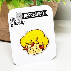May include: A die-cut sticker featuring a cartoon girl with yellow hair and a smiling face. The sticker is on a white card with the text "Our Girl Ghosty" and "Refreshed".