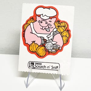 May include: A vintage "Scratch n' Sniff" sticker featuring a cartoon pig chef wearing a white hat and apron, surrounded by yellow onions. The pig is pink with black outlines, set against a red border. The sticker has the "TREND" logo.