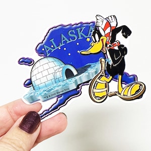 May include: A blue and white magnet shaped like the state of Alaska with a cartoon duck wearing a red and white scarf and snowshoes. The magnet says "Alaska" and has a small igloo on it. The text "TM & © 2000 Warner Bros." is at the bottom of the magnet.