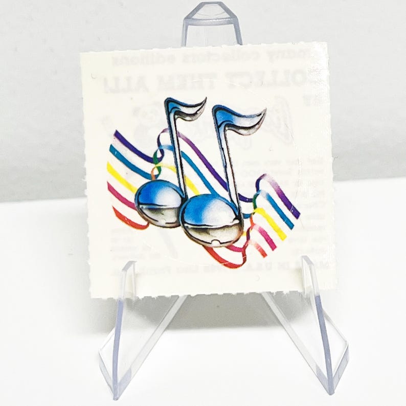 May include: A square, off-white sticker featuring two blue musical notes with silver accents and rainbow-colored streamers. The sticker is displayed on a clear plastic stand.