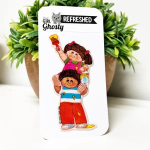 Puede incluir: Una pegatina blanca con una ilustración de dibujos animados de dos niños. Uno está sobre los hombros del otro, sosteniendo un pincel y un bote de pintura. La pegatina tiene el texto "Our Girl Ghosty REFRESHED" y "© 1983 OAA INC."