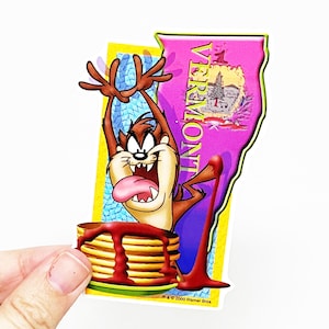 May include: A sticker featuring a cartoon Tasmanian Devil character, with a wide grin and tongue sticking out, holding a stack of pancakes with syrup dripping down the side. The sticker is shaped like the state of Vermont, with the word "VERMONT" written vertically down the side. The sticker has a yellow border and the text "TM & © 2000 Warner Bros."