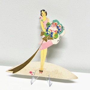 Könnte beinhalten: Vintage Papierauschnitt einer Frau in einem rosa Kleid, die einen großen Blumenstrauß hält. Der Ausschnitt hat ein goldenes Band und eine lange goldene Schleppe. Die Frau steht auf einer beigen Basis mit einem transparenten Kunststoffständer.