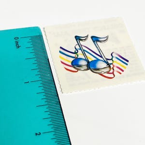 May include: A postage stamp featuring two blue musical notes with rainbow-colored ribbons. The stamp is approximately 1 inch in height. The background is white, and the stamp has a perforated edge.