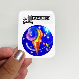 May include: A sticker with an ice cream cone illustration in vibrant colours. The ice cream is a blend of purple, yellow, and orange, set against a blue backdrop with stars and planets. The sticker also includes the text "Our Girl Ghosty" and "Refreshed".